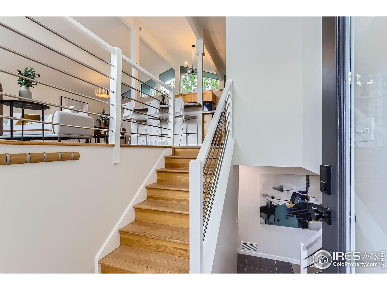 This interior shot showcases a modern hallway and staircase. The staircase features light wood steps and a sleek railing with horizontal metal bars. A piece of abstract art hangs on the wall beneath the stairs, adding a touch of contemporary style to the space.