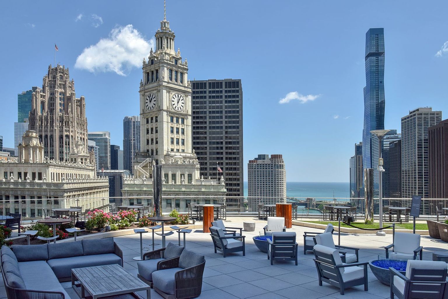 This expansive rooftop terrace offers a sophisticated outdoor lounge area with comfortable grey seating, modern fire pits, and manicured planters. The space provides a dramatic, cinematic perspective of the Chicago skyline, featuring iconic architecture like the Wrigley Building clock tower and the St. Regis Chicago. The open-air setting creates an inviting atmosphere for entertaining against a backdrop of urban grandeur.
