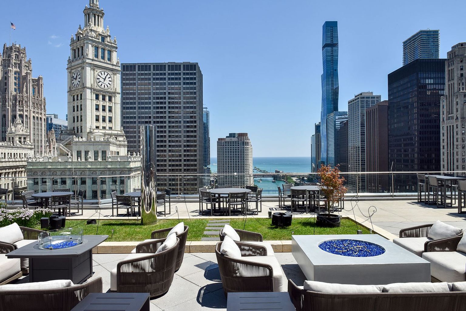 This expansive rooftop terrace offers a sophisticated outdoor living space with panoramic views of the Chicago skyline, including the iconic Wrigley Building clock tower. The area is furnished with multiple seating arrangements, including comfortable lounge chairs surrounding two modern fire pits with blue glass accents and several dining tables. The setting combines urban luxury with open-air comfort, providing an ideal vantage point for city living.