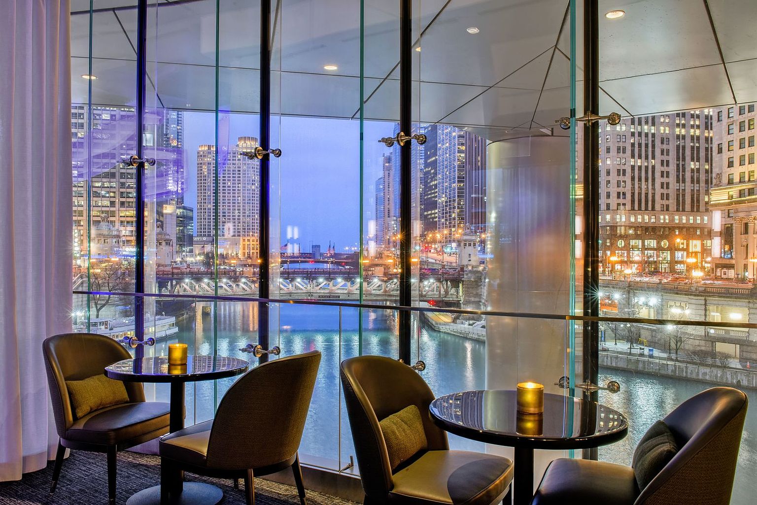 This sophisticated lounge area features floor-to-ceiling glass walls that provide a panoramic view of a city riverfront and illuminated urban skyline. The space is furnished with dark, modern armchairs and small circular tables, each accented by a warm, glowing candle. The atmosphere is elegant and serene, highlighting the property's prime location and high-end interior design.