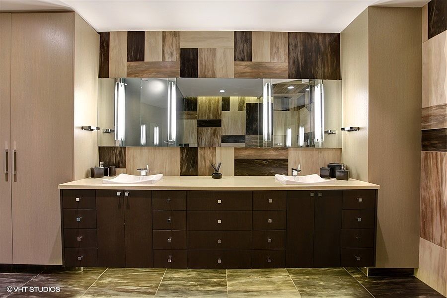 This primary bathroom features a double vanity with a dark wood cabinet base and a light-toned countertop. The wall behind the vanity is adorned with a decorative, multi-toned tile mosaic that extends behind the mirrors, creating a sophisticated focal point. The space is illuminated by integrated vertical lighting on the mirrors and recessed ceiling lights, providing a clean and contemporary aesthetic.