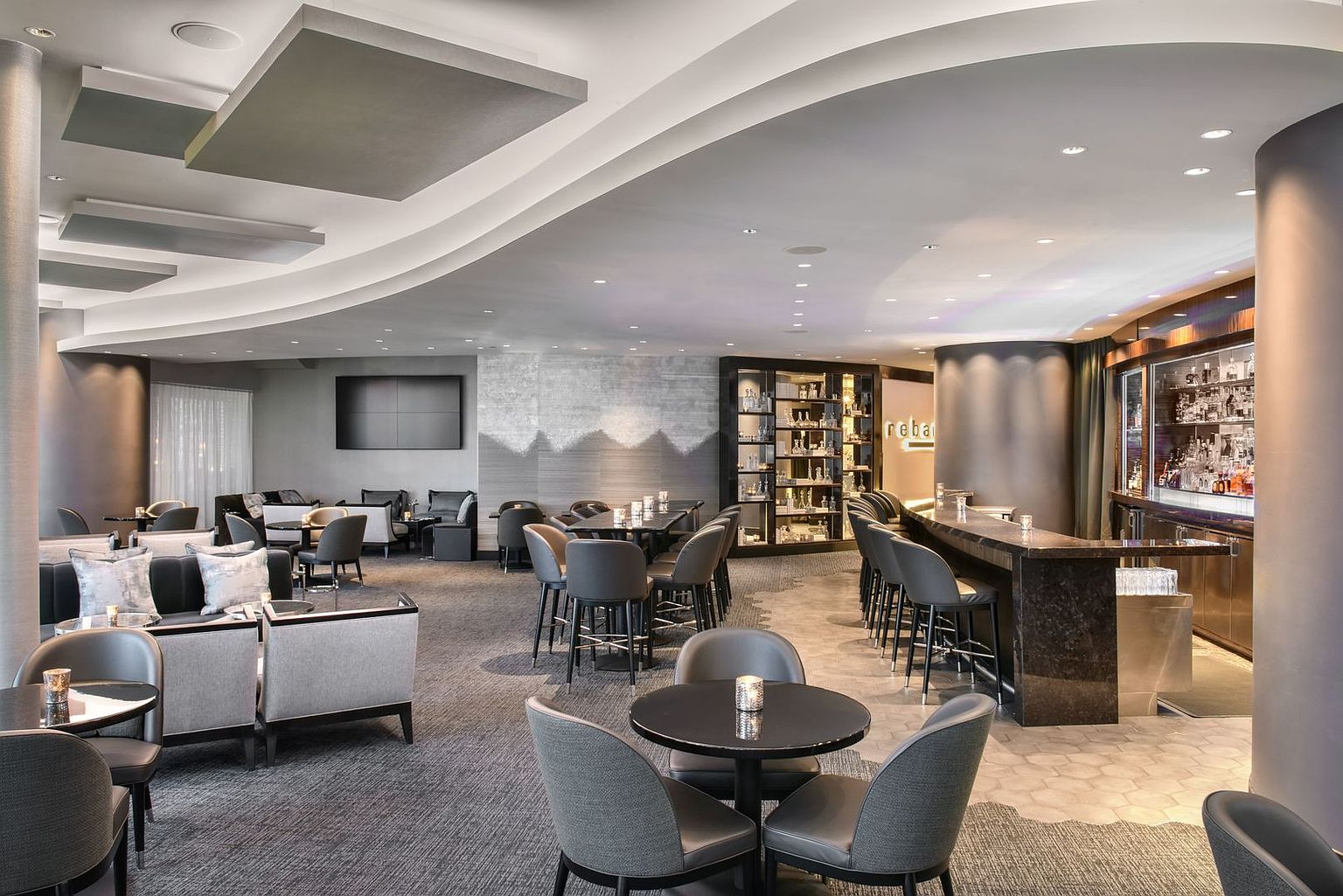 This sophisticated lounge and bar area features a contemporary design with a mix of plush gray seating, dark circular tables, and a prominent stone-topped bar. The space is illuminated by recessed lighting and elegant fixtures, creating an inviting atmosphere for social gatherings. A large, wall-mounted television and a glass-fronted display cabinet add functional and aesthetic appeal to this upscale communal amenity.