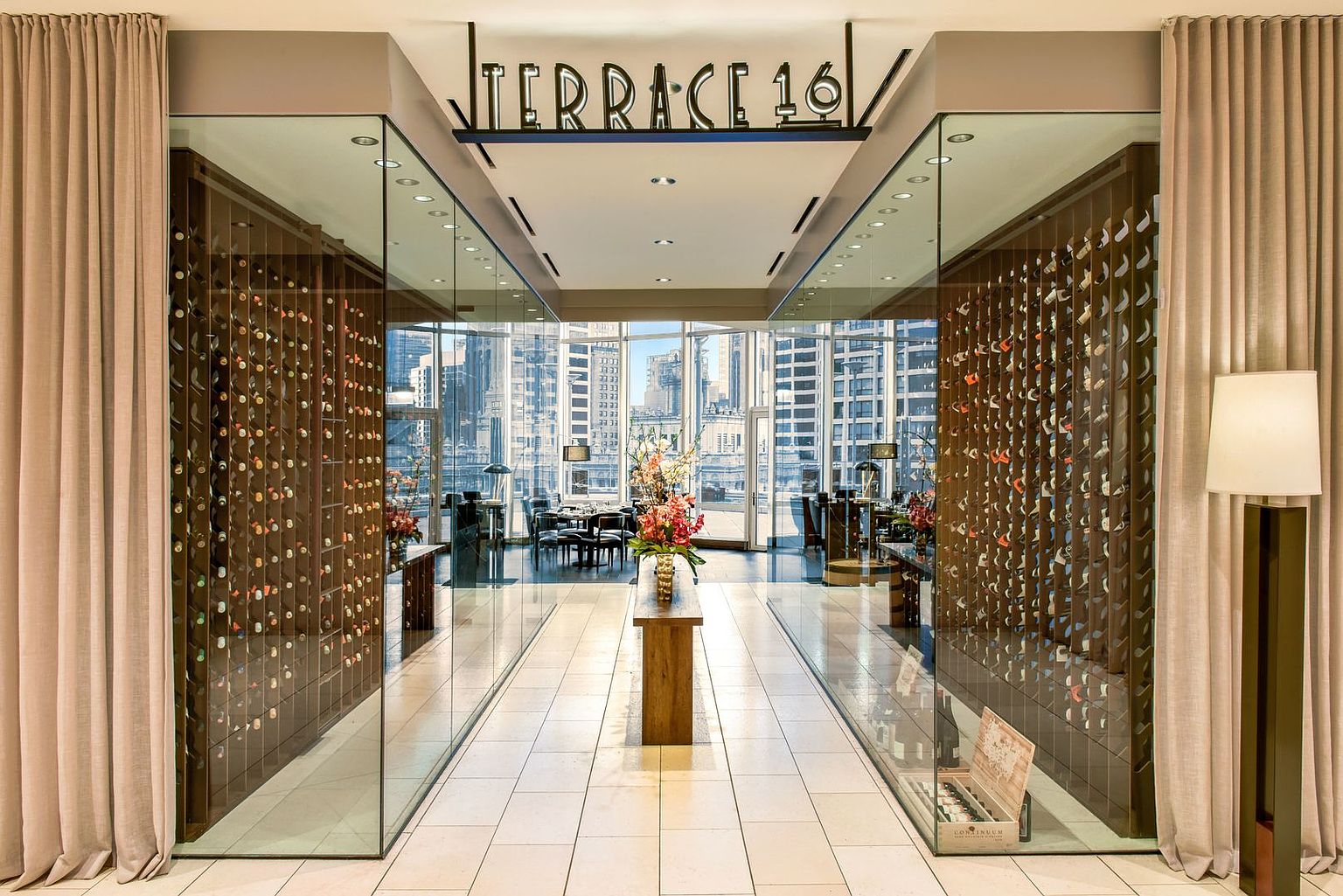 This sophisticated entryway leads into a restaurant space, framed by two expansive, glass-enclosed wine storage displays that showcase extensive bottle collections. The central path features a sleek wooden console table topped with a vibrant floral arrangement, drawing the eye toward the bright, open dining area beyond. The space is defined by a clean, modern aesthetic with neutral tones, light-colored tile flooring, and soft drapery flanking the entrance.