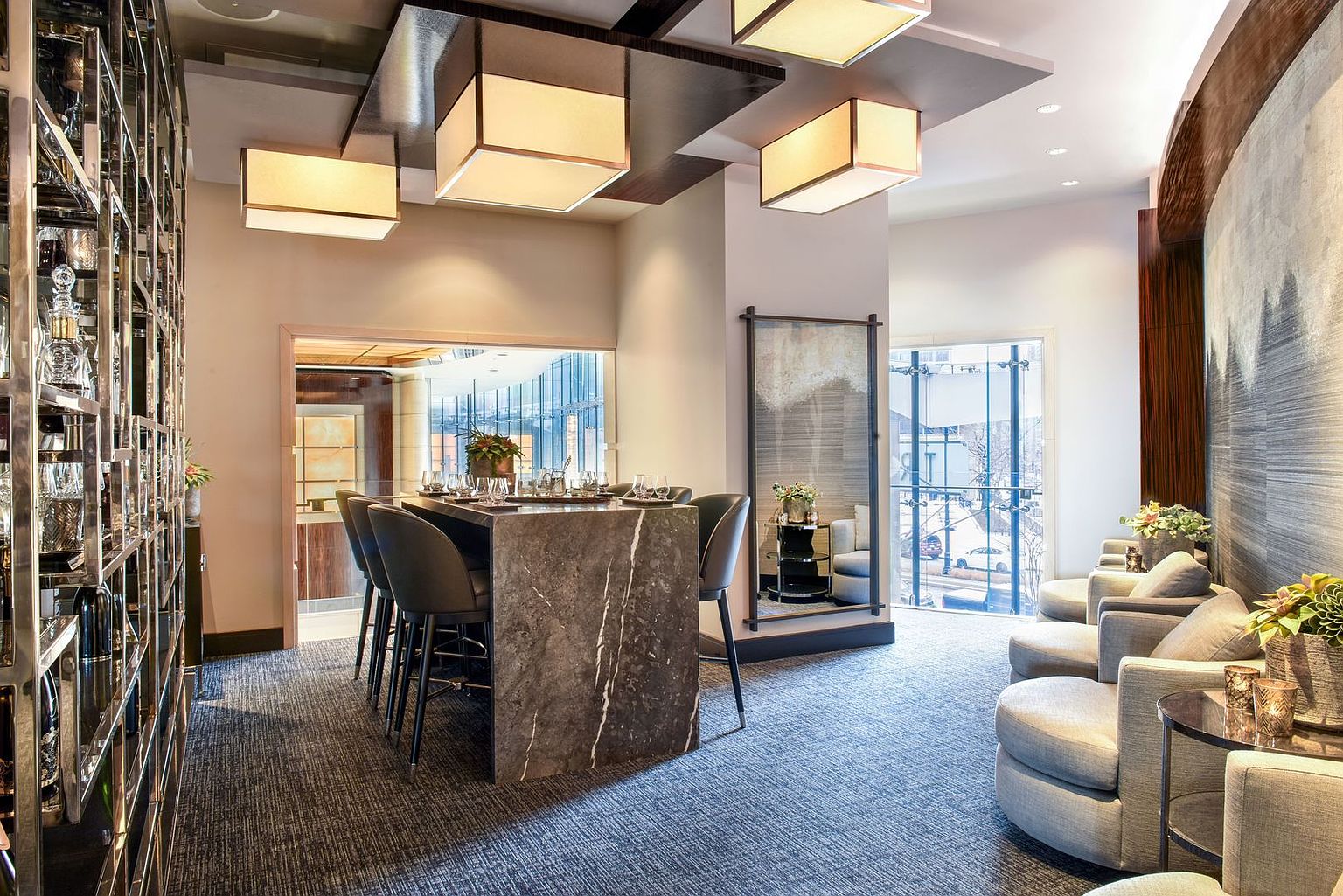 This sophisticated lounge area features a central stone-topped bar table surrounded by dark upholstered stools, illuminated by modern geometric pendant lights. To the left, a tall, multi-tiered metal shelving unit displays glassware, while the right side offers a cozy seating arrangement with plush armchairs and a small side table. The space is framed by large windows that provide a view of the street, creating an elegant and inviting atmosphere for social gatherings.
