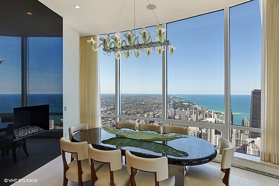 This sophisticated dining room features a dark, polished oval table surrounded by eight cream-colored upholstered chairs. A unique, multi-armed glass chandelier hangs above the table, while the room is framed by expansive floor-to-ceiling windows that offer a panoramic view of the city skyline and the lake. To the left, a sleek, dark-toned wall incorporates a modern fireplace, creating a dramatic contrast against the bright, airy atmosphere of the space.