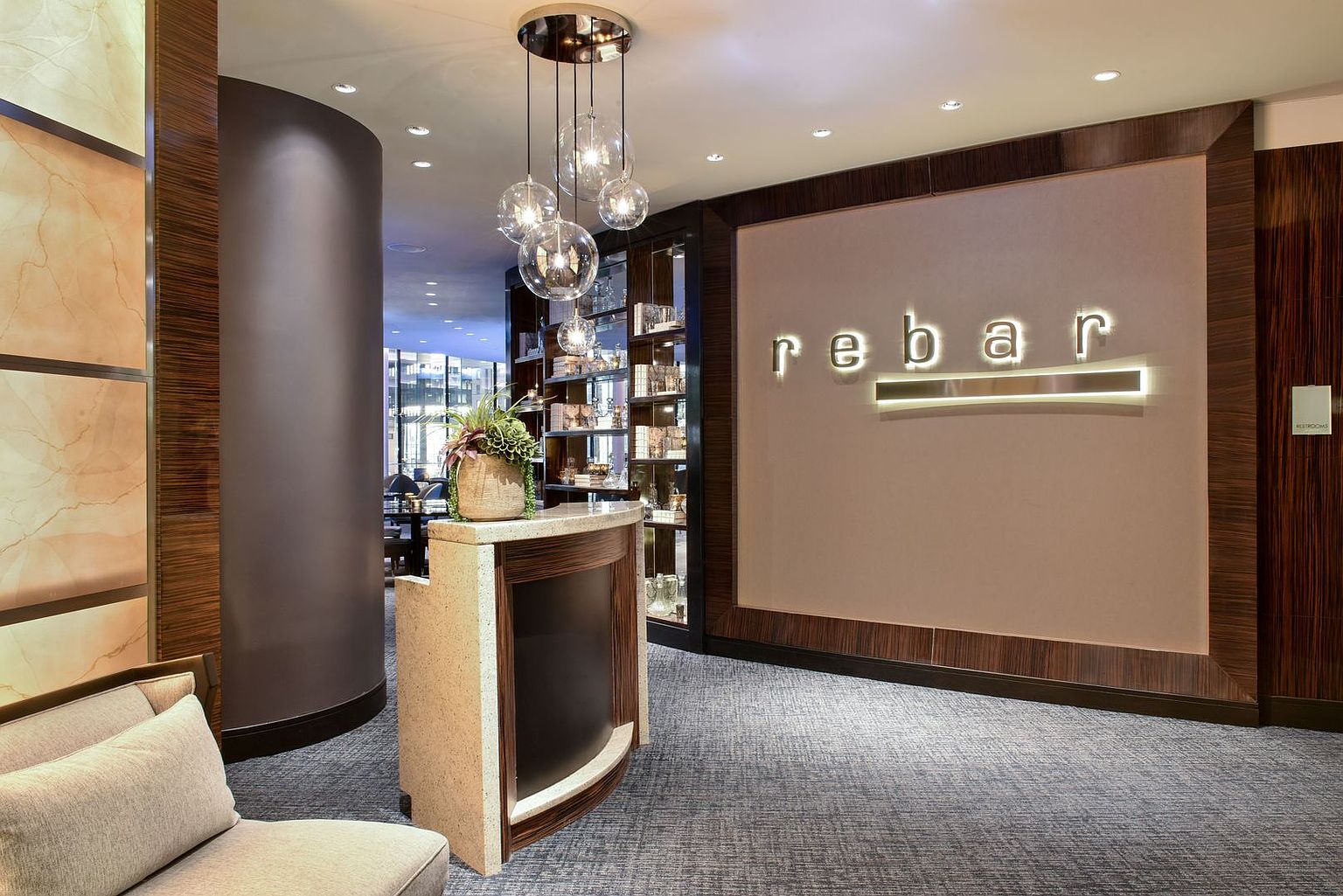 This image captures the sophisticated entrance of a commercial space named 'rebar,' featuring a sleek reception desk and a prominent wall-mounted sign. The interior design utilizes a mix of dark wood paneling, warm ambient lighting, and a cluster of glass globe pendant lights to create an upscale, welcoming atmosphere. A small display shelf with glassware and a potted plant on the desk add refined decorative touches to the professional lobby area.