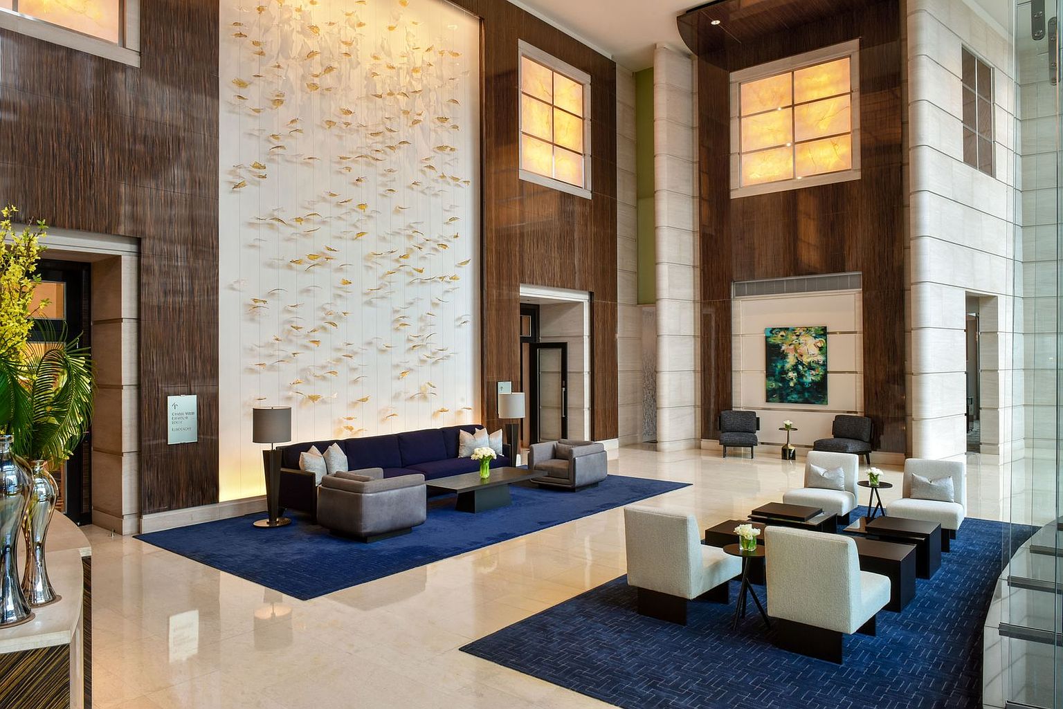 This grand, high-ceilinged lobby features a sophisticated seating area arranged on a deep blue rug, anchored by a dark sofa and matching armchairs. A striking, large-scale wall installation of delicate, leaf-like accents serves as a focal point, complemented by warm, glowing light panels set into the dark wood-paneled walls. The polished stone flooring and symmetrical layout create an atmosphere of refined luxury and professional elegance.