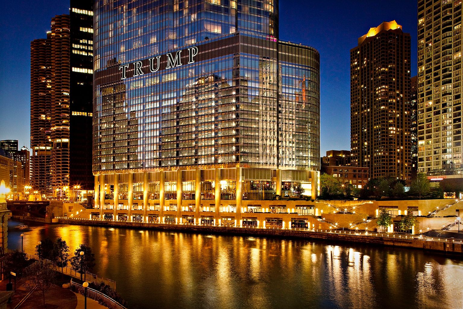 This striking night-time shot captures the iconic Trump International Hotel & Tower in Chicago, prominently displaying the 'TRUMP' signage on its glass-clad facade. The building's modern architectural design is highlighted by warm, golden interior lighting that reflects beautifully onto the calm surface of the Chicago River below. The perspective is a cinematic, eye-level view from across the water, showcasing the skyscraper's imposing scale amidst the surrounding urban skyline.