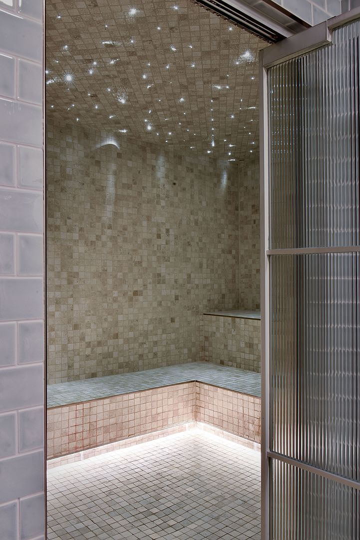 This image captures a luxurious steam shower or sauna featuring a mosaic-tiled interior with a dramatic fiber-optic starlight ceiling. The space includes built-in tiled seating and is accessed through a modern, ribbed glass door. Soft, integrated under-bench lighting creates a serene and spa-like atmosphere, highlighting the intricate tile work.