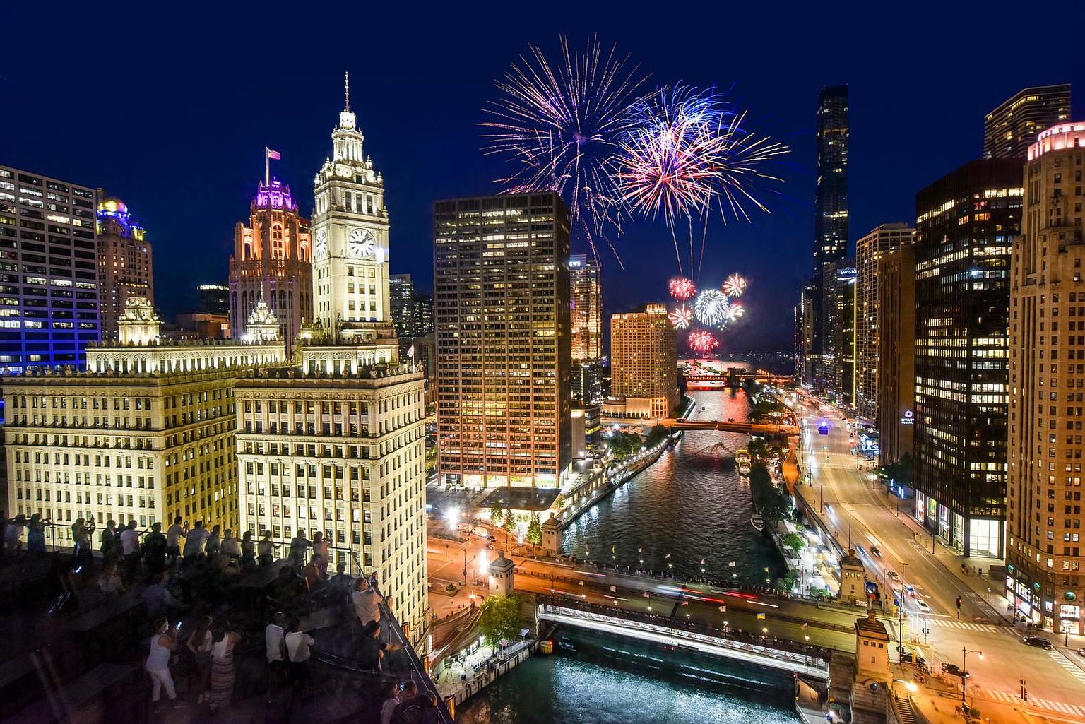 This high-angle aerial view captures a vibrant cityscape at night, featuring the iconic Wrigley Building and surrounding Chicago architecture illuminated against a deep blue sky. Fireworks burst above the river, which winds through the dense urban landscape, reflecting the city lights and bridge illumination. In the foreground, a rooftop terrace is occupied by people enjoying the panoramic view, emphasizing the property's prime location for entertainment and city living.