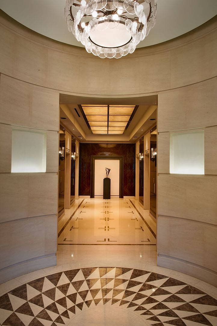 This elegant, symmetrical hallway features a grand circular foyer with a patterned stone floor leading into a long, illuminated corridor. The space is defined by curved, light-toned stone walls, a sophisticated tiered chandelier, and a central sculpture positioned at the end of the hall. The perspective is centered and cinematic, emphasizing the high-end architectural design and the warm, ambient lighting throughout the passage.