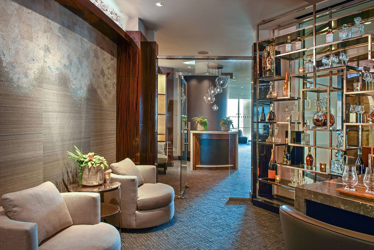 This sophisticated lounge area features a refined seating arrangement with two plush, neutral-toned armchairs positioned near a small side table. To the right, a striking, multi-tiered metal shelving unit displays an array of glassware and spirits, while a glass partition leads to a secondary reception area with a curved desk and modern pendant lighting. The space exudes a high-end, professional atmosphere, characterized by textured wall coverings and a polished, contemporary aesthetic.