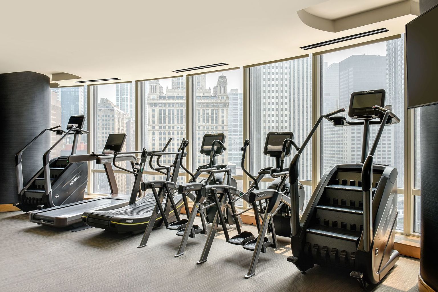 This modern, well-equipped home gym features a row of professional-grade fitness machines, including treadmills and stair climbers, arranged neatly on a neutral-toned floor. The space is defined by expansive, floor-to-ceiling windows that provide a panoramic view of the surrounding city skyline, filling the room with natural light. The clean, minimalist aesthetic and high-end equipment create a sophisticated and motivating environment for exercise.