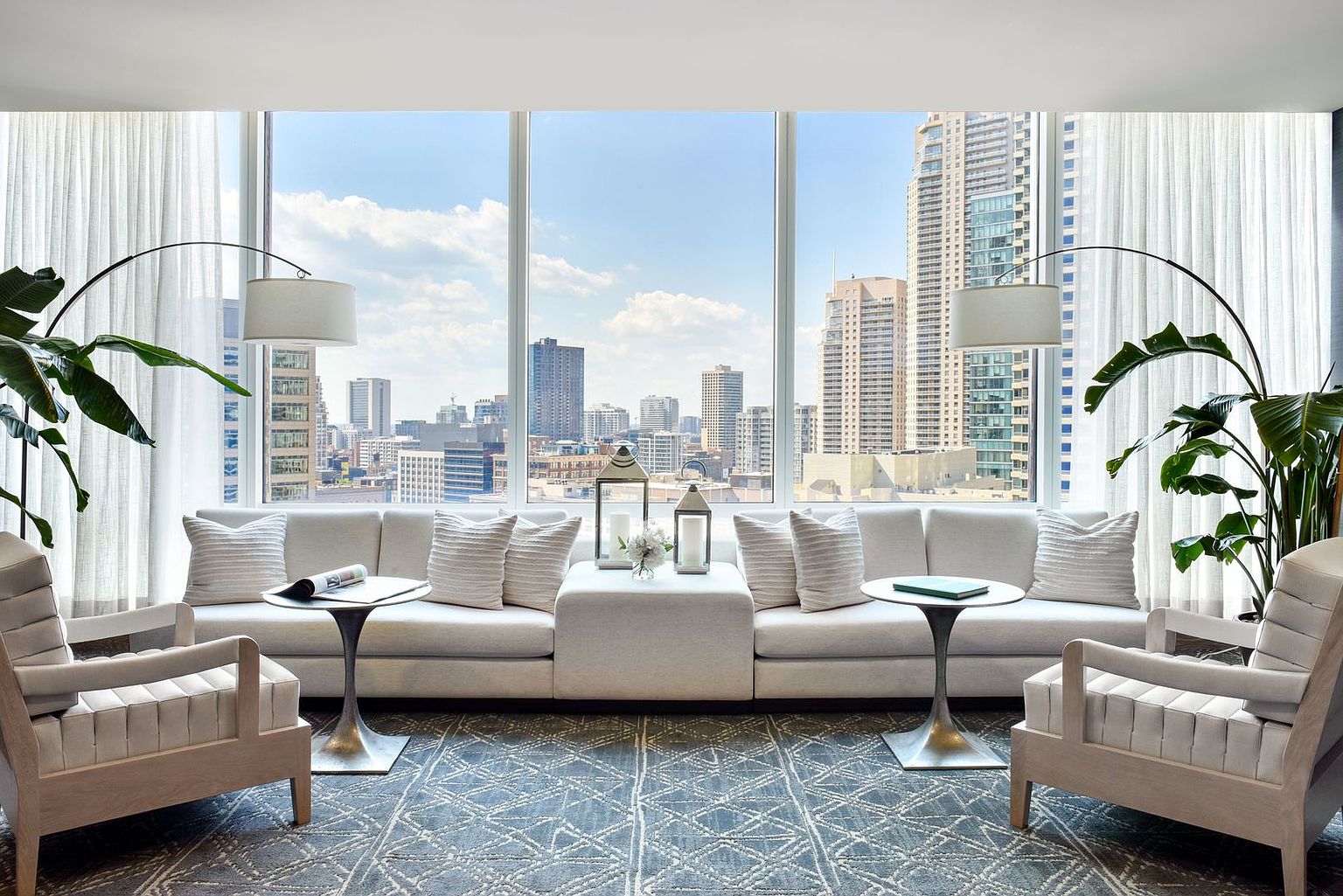 This sophisticated living room features a symmetrical layout centered around a long, light-toned sofa and two matching armchairs. Large, floor-to-ceiling windows provide an expansive, unobstructed view of the city skyline, bathing the room in natural light. The space is accented by two tall, arched floor lamps, potted plants, and a patterned area rug, creating a refined and airy urban atmosphere.