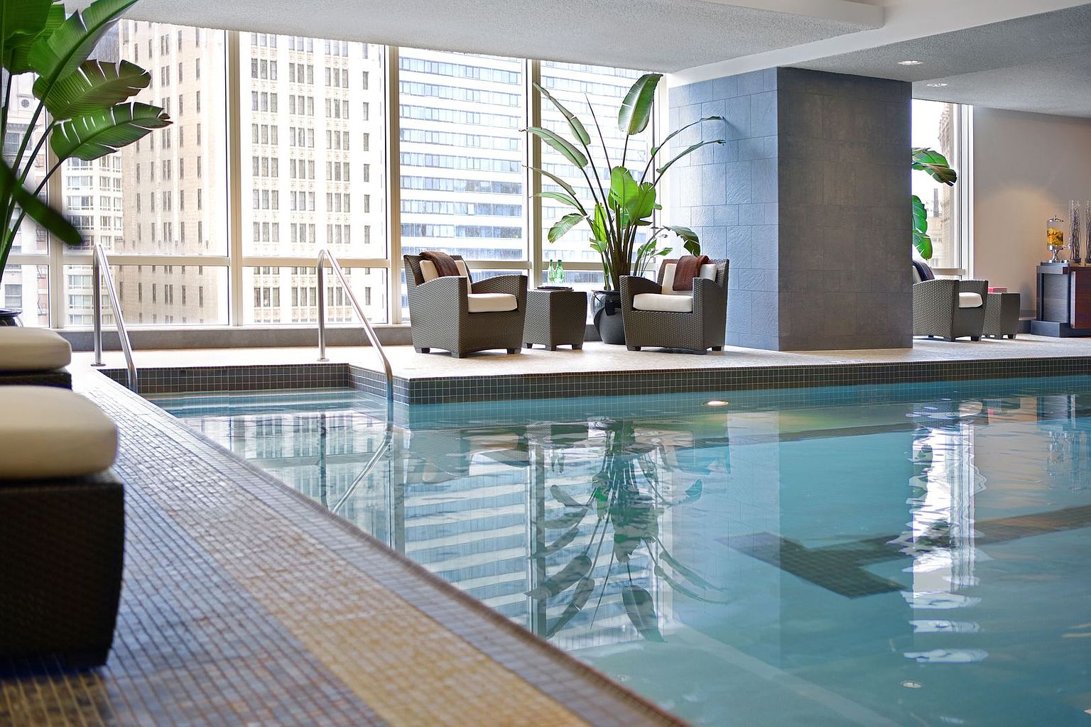 This indoor pool area features a serene, modern aesthetic with large windows offering expansive city views. The space includes a tiled pool, a raised deck area with comfortable lounge seating, and lush potted plants that enhance the tranquil atmosphere. The perspective is from the poolside, capturing the reflection of the interior and the urban skyline on the water's surface.