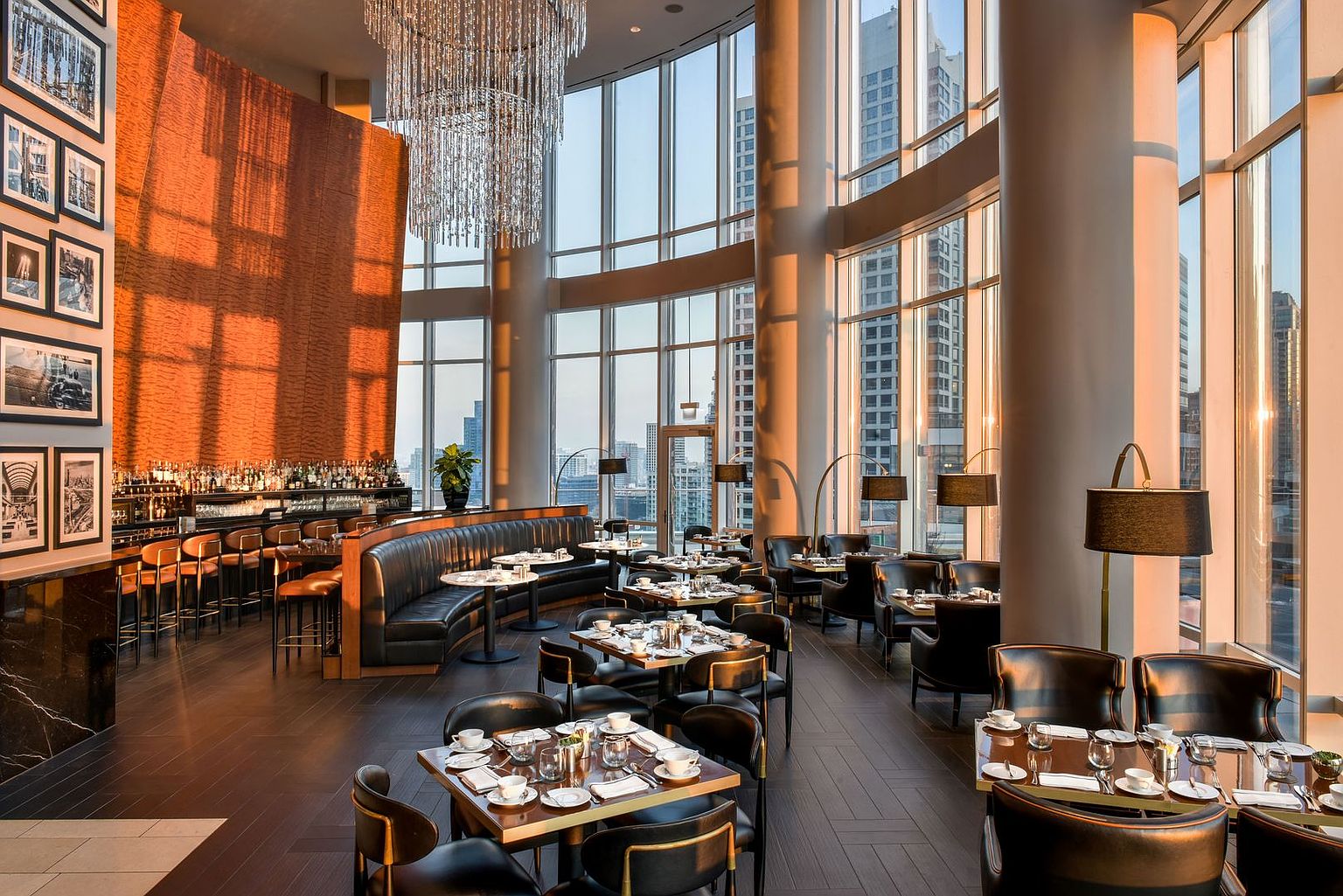 This sophisticated restaurant dining area features a high-ceilinged space with expansive floor-to-ceiling windows that offer panoramic city views. The room is anchored by a large, curved leather banquette and a striking, textured copper-toned accent wall, complemented by a dramatic crystal chandelier. The layout includes multiple dining tables set with white linens and dark leather chairs, creating an upscale, modern atmosphere perfect for fine dining.