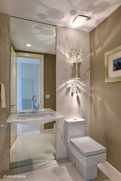 This elegant powder room features a wall-mounted floating sink and a modern, one-piece toilet set against a striking stone-clad wall. A decorative, multi-armed wall sconce with crystal accents provides warm lighting, while a large mirror reflects the adjacent hallway. The space is finished with neutral, textured wall coverings and light-toned flooring, creating a sophisticated and compact atmosphere.
