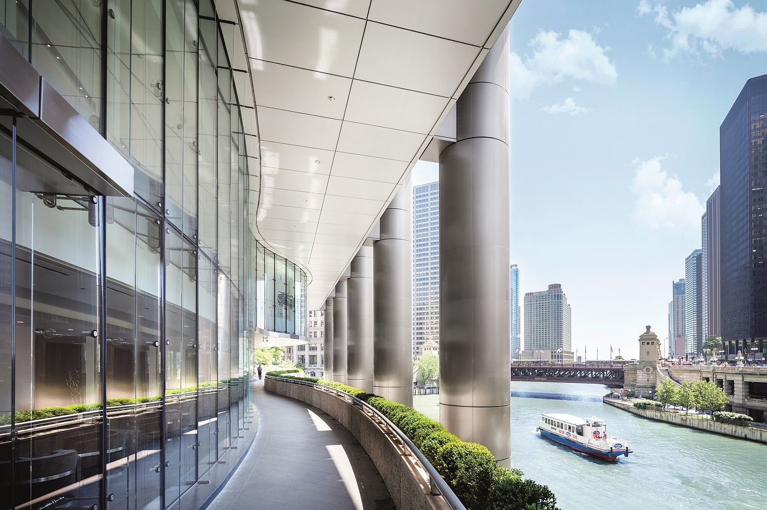 This image captures a sleek, modern walkway along the riverfront, characterized by a curved glass facade and substantial metallic columns. The path is lined with manicured hedges and offers a direct, scenic view of the river, a passing water taxi, and the surrounding urban skyline. The perspective emphasizes the architectural integration of the building with the waterfront, creating a sophisticated and airy atmosphere.
