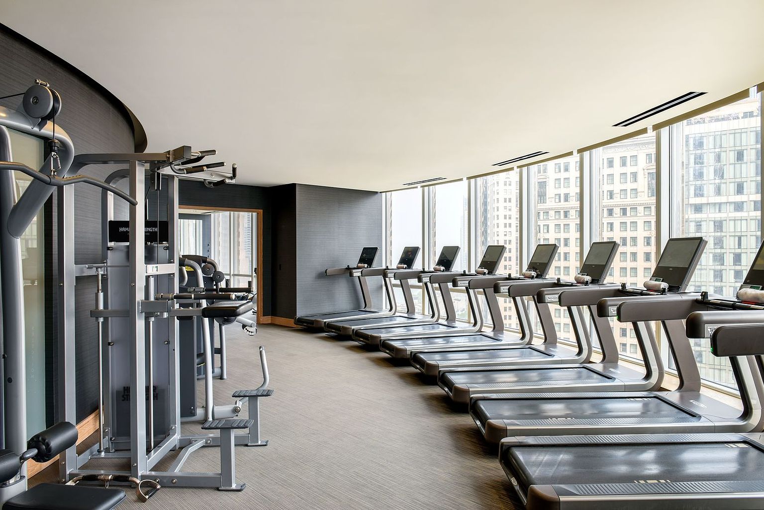 This modern, high-end fitness center features a row of professional-grade treadmills positioned alongside a wall of floor-to-ceiling windows that offer expansive city views. To the left, a multi-station strength training machine provides a comprehensive workout area, all set against dark, textured walls and neutral carpeting. The space is bright and airy, conveying a clean, sophisticated atmosphere suitable for a luxury residential building.