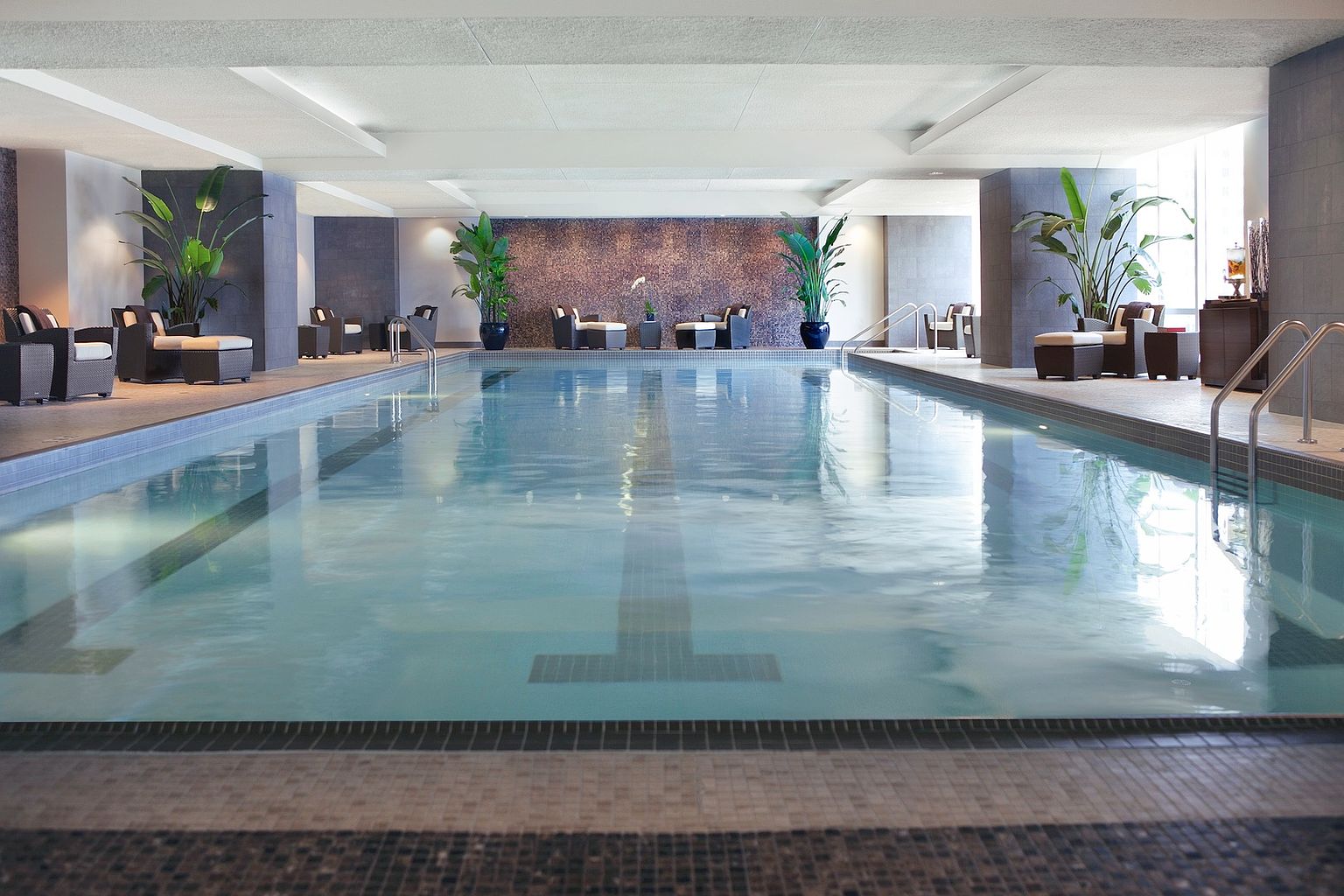 This indoor swimming pool area features a long, rectangular lap pool as the central focus, framed by dark stone pillars and a textured, mosaic-style accent wall at the far end. The space is furnished with several seating areas consisting of cushioned armchairs and ottomans, complemented by large potted plants that add a touch of greenery. The perspective is a low-angle, symmetrical shot from the edge of the pool, emphasizing the clean lines and tranquil, upscale atmosphere of the facility.