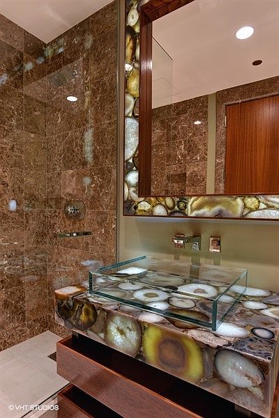 This luxurious bathroom features a striking vanity countertop and mirror frame crafted from backlit, multi-colored agate stone. A clear glass vessel sink sits atop the illuminated vanity, which is paired with dark wood cabinetry and a walk-in shower enclosed by glass. The walls are finished in large, polished brown stone tiles, creating a sophisticated and high-end aesthetic.