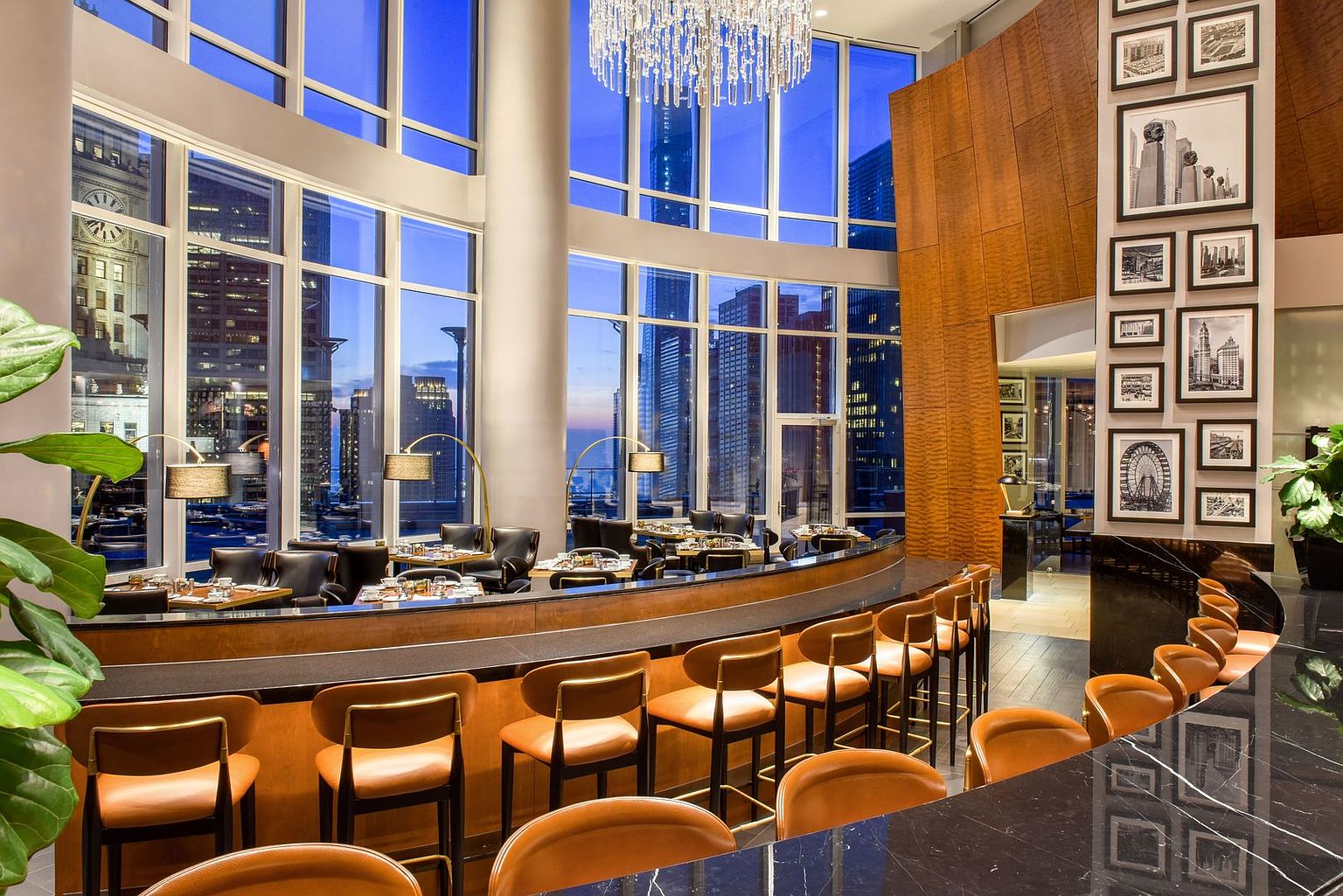 This sophisticated lounge or restaurant space features a curved bar with leather-upholstered stools, set against a backdrop of expansive floor-to-ceiling windows that offer a panoramic view of the city skyline at dusk. A prominent wood-paneled wall displays a curated gallery of framed black-and-white architectural photographs, while a large, multi-tiered crystal chandelier hangs from the high ceiling. The atmosphere is upscale and modern, designed to provide a luxurious setting for social gatherings.