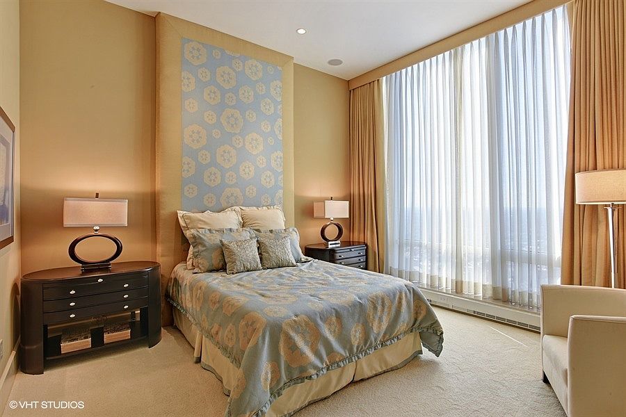 This elegant bedroom features a central bed with a patterned blue and beige comforter, set against a decorative, light-blue patterned accent wall. To the left, a dark wood dresser with a matching lamp provides storage, while a large window with floor-to-ceiling sheer curtains allows ample natural light to fill the room. A comfortable armchair sits in the corner, completing the sophisticated and serene atmosphere of this well-appointed space.