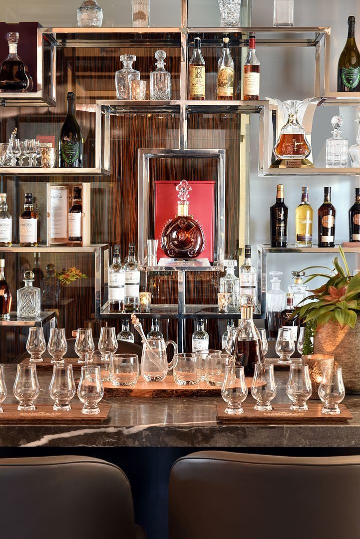 This image showcases a sophisticated home bar area featuring an intricate, multi-tiered metal shelving unit filled with an extensive collection of premium spirits and crystal decanters. A dark stone countertop serves as the bar surface, neatly arranged with numerous tasting glasses and a central pitcher, creating an inviting atmosphere for entertaining. The backdrop consists of rich, dark wood paneling that adds warmth and elegance to the refined, upscale aesthetic of the space.