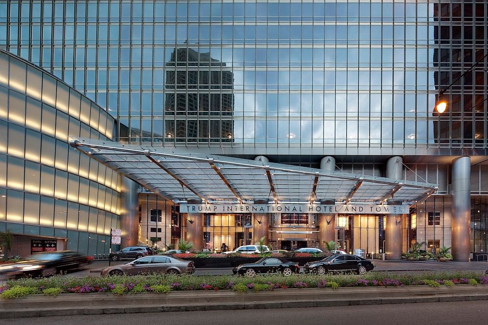 This image captures the grand, modern entryway of the Trump International Hotel and Tower, characterized by a massive, sleek glass canopy extending over the drop-off area. The building's facade features expansive, reflective glass panels that mirror the surrounding urban architecture, creating a sophisticated and imposing aesthetic. Several luxury vehicles are positioned in the driveway, emphasizing the upscale nature of the property, while a manicured garden bed with vibrant flowers adds a touch of color to the polished metallic and glass environment.
