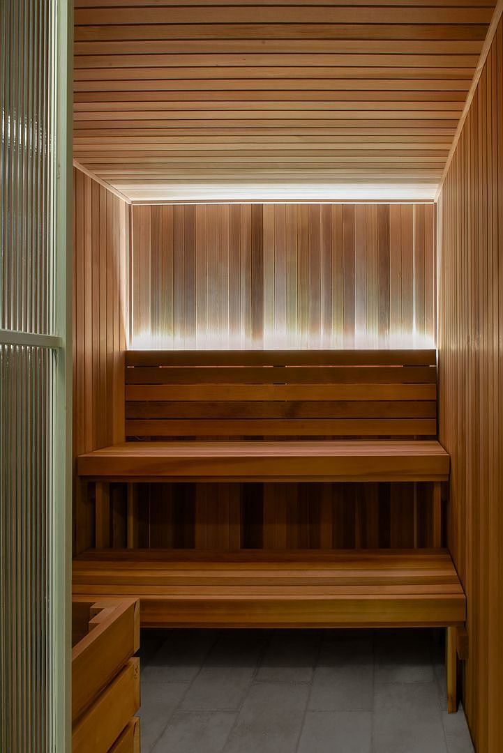 This image captures the interior of a modern sauna, characterized by warm-toned wood paneling on the walls and ceiling. Two tiered benches provide seating, while a backlit, textured glass panel at the back creates a soft, diffused glow. The perspective is a straight-on view from the doorway, emphasizing the clean lines and minimalist design of this wellness space.