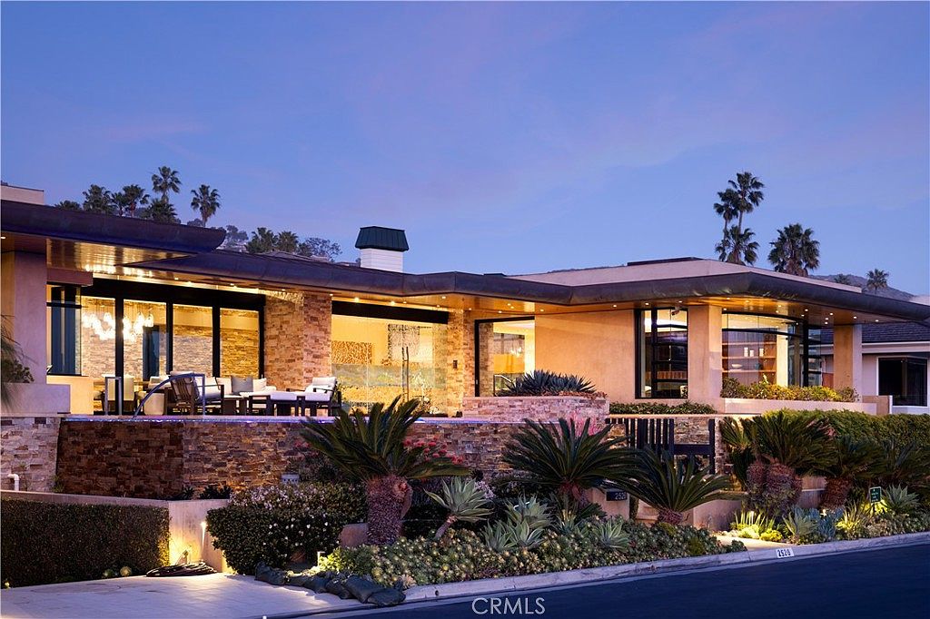 This is a front exterior view of a modern luxury home at dusk. The house features a combination of stone and stucco facade, large windows, and a flat roof with an overhang. Lush landscaping with palm trees and various plants adds to the property's curb appeal, creating an inviting and upscale impression.