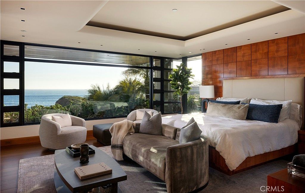 This is a luxurious primary bedroom with a stunning ocean view through large windows. The room features a king-size bed with plush bedding, a seating area with a stylish chaise lounge, and rich wood paneling on the wall behind the bed. The overall impression is one of elegance and tranquility, perfect for relaxation.