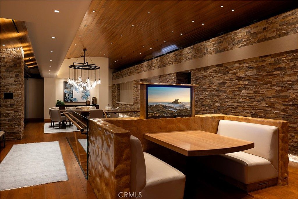 This interior shot showcases a dining area with a unique built-in booth seating arrangement, complemented by a wooden table. A modern chandelier hangs above a separate dining table in the background, and the room features a stone accent wall and wood-paneled ceiling, creating a warm and inviting atmosphere. A television is mounted on the booth structure.
