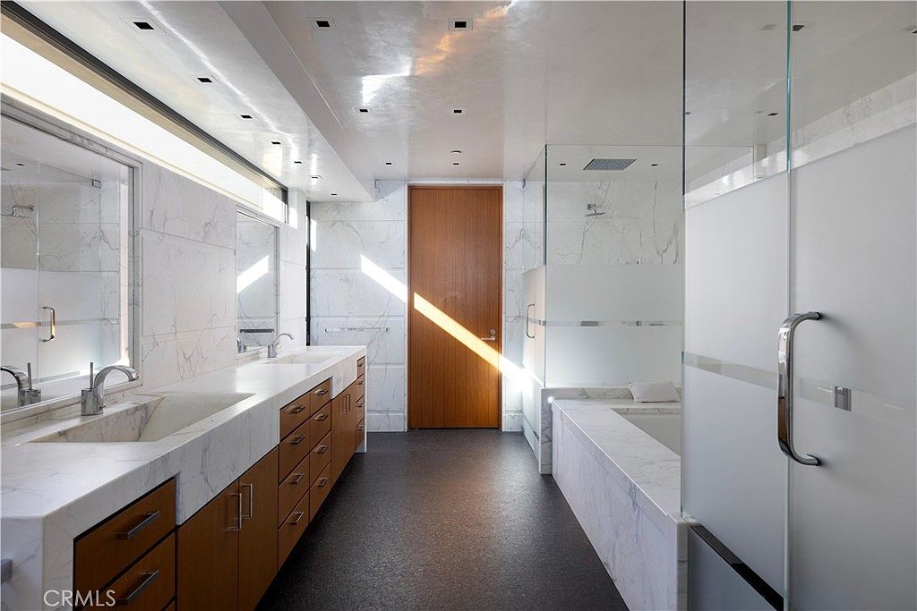This is a modern primary bathroom featuring a double vanity with marble countertops and wood cabinetry. The bathroom includes a glass-enclosed shower and a separate bathtub area, both with marble walls. The flooring is a dark, contrasting material, and a wooden door is visible in the center, adding warmth to the space.