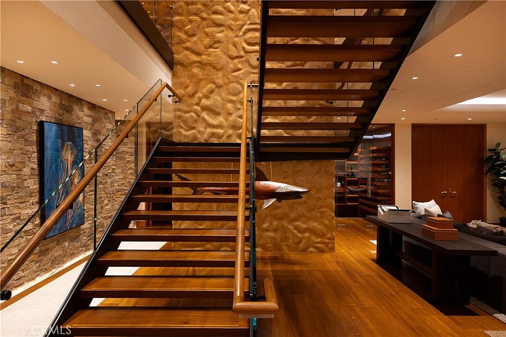 This interior shot showcases a modern hallway with striking architectural details. The wooden staircase features glass railings and a unique shark sculpture mounted on the wall. The hardwood flooring and textured wall finishes add warmth and sophistication to the space, creating a luxurious and inviting atmosphere.