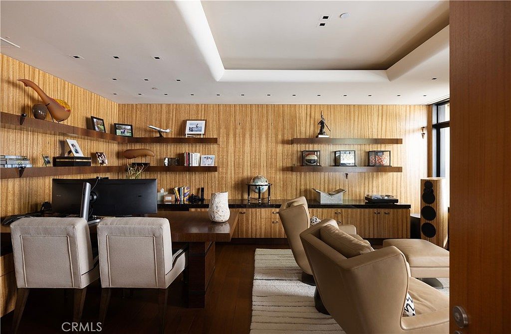 This is an interior shot of a well-appointed office or study. The room features wood-paneled walls, built-in shelving displaying various decorative items, and a large desk with two chairs. A seating area with comfortable armchairs and a rug adds a touch of relaxation to the workspace.