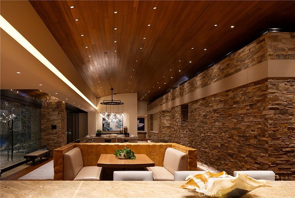 This interior shot showcases a dining area with a unique built-in booth seating arrangement around a wooden table, complemented by modern chairs. The room features a striking stone wall and a warm wood plank ceiling with recessed lighting, creating a sophisticated and inviting atmosphere. A chandelier hangs elegantly in the background, adding to the room's upscale aesthetic.