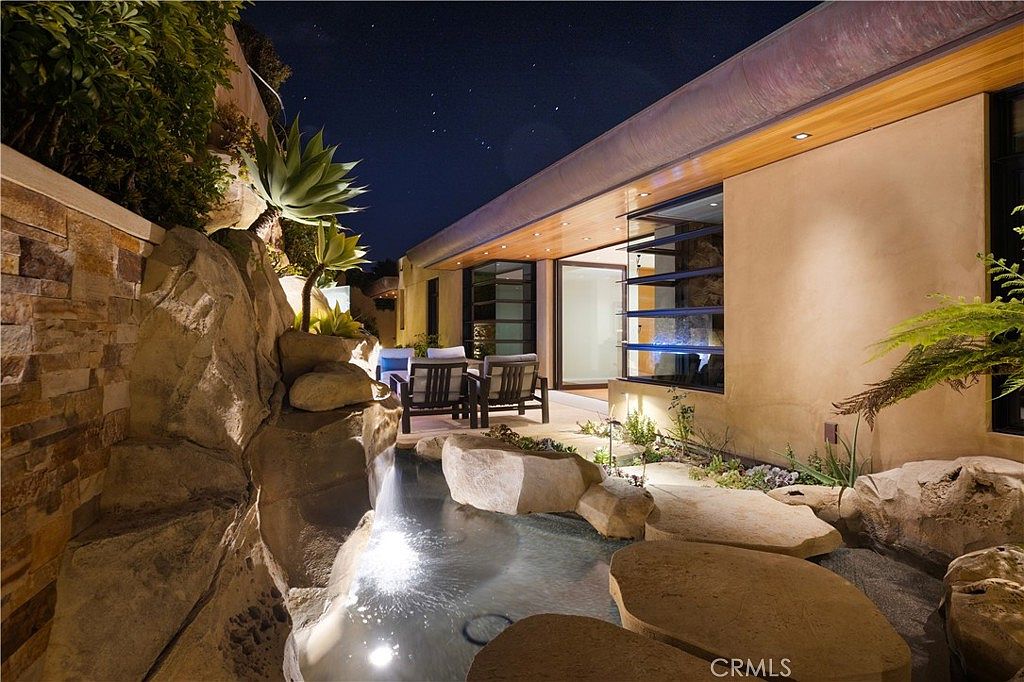 This image showcases a luxurious outdoor pool and spa area at night. The pool features a naturalistic design with large rocks and a small waterfall, creating a serene and inviting atmosphere. The adjacent building has modern architectural elements, including large windows and a sleek roofline, enhancing the overall appeal of the property.