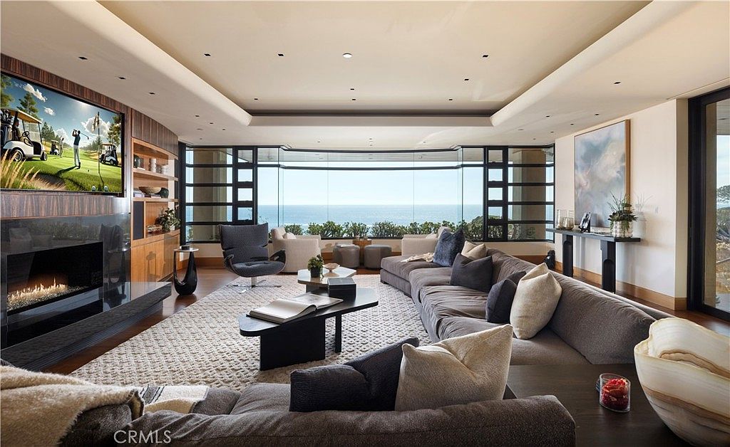 This is a luxurious living room featuring a large sectional sofa, a modern fireplace with a TV above, and floor-to-ceiling windows offering ocean views. The room is decorated in neutral tones with pops of color from the artwork and accessories, creating a sophisticated and inviting atmosphere. The perspective is from the front of the room, looking towards the ocean view.