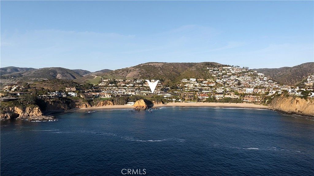 This aerial shot showcases a coastal community nestled along a picturesque bay. The scene features a mix of residential homes and natural landscapes, with the ocean stretching out to the horizon under a clear sky. The perspective highlights the proximity of the homes to the water and the overall serene setting, ideal for potential buyers seeking a coastal lifestyle.