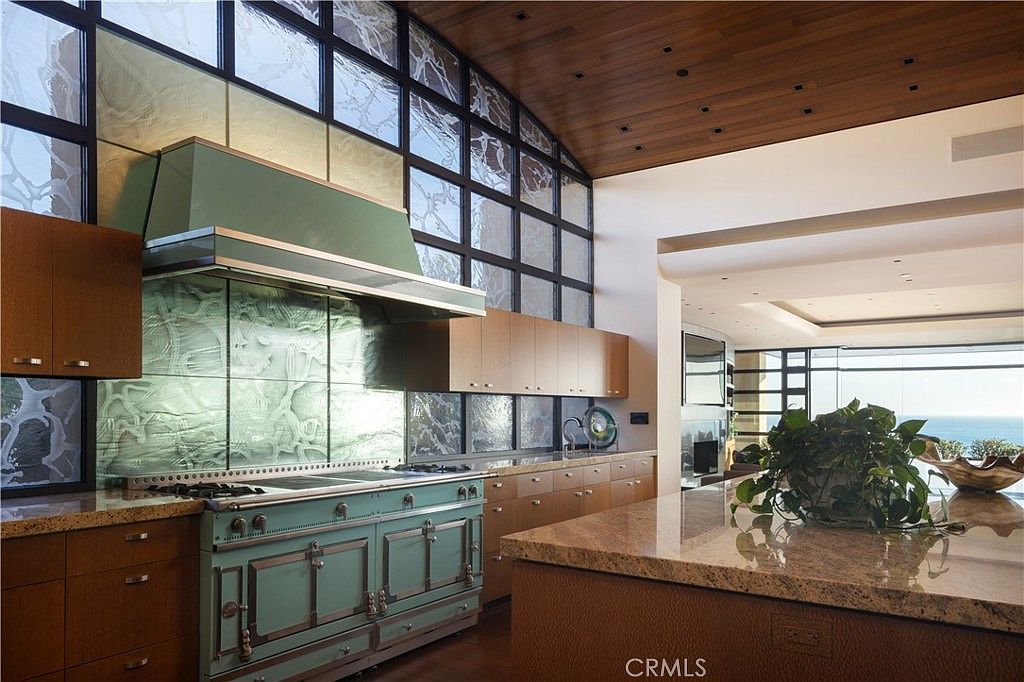This is a well-lit kitchen featuring custom cabinetry, a large island with a granite countertop, and a high-end range with a custom hood. The kitchen has a modern design with a large window that provides natural light and a view. The overall impression is luxurious and spacious.