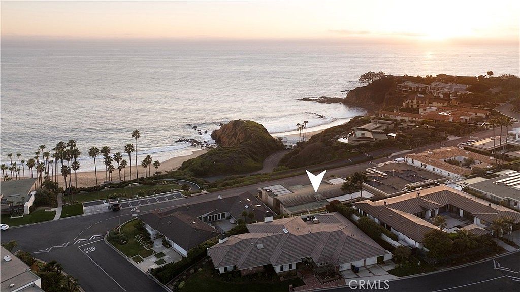 This aerial shot showcases a luxurious coastal property with a sprawling tile roof, surrounded by lush landscaping and neighboring homes. The property is situated near a picturesque beach with palm trees and a rocky coastline, offering stunning ocean views. The overall impression is one of upscale coastal living and serene beauty.