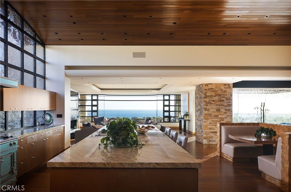This is a spacious, open-concept kitchen and dining area with a modern design. The kitchen features wooden cabinets, granite countertops, and a large island with seating. The dining area includes a booth and table, with large windows offering ocean views, creating a luxurious and inviting atmosphere.