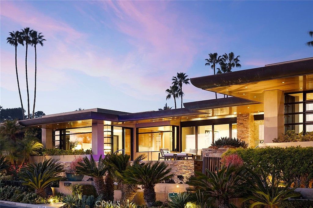 This is a front exterior view of a modern luxury home at dusk. The architecture features clean lines, large windows, and a flat roof with overhangs. Lush landscaping with palm trees and other tropical plants surrounds the property, enhancing its curb appeal and creating a serene atmosphere.