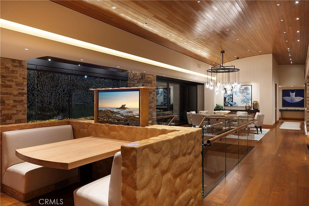 This is an interior shot of a dining area featuring a built-in booth with a wooden table and a TV. The dining area includes a modern chandelier, a dining table with chairs, and artwork on the walls. The space is well-lit with a wooden ceiling and hardwood floors, creating a warm and inviting atmosphere.