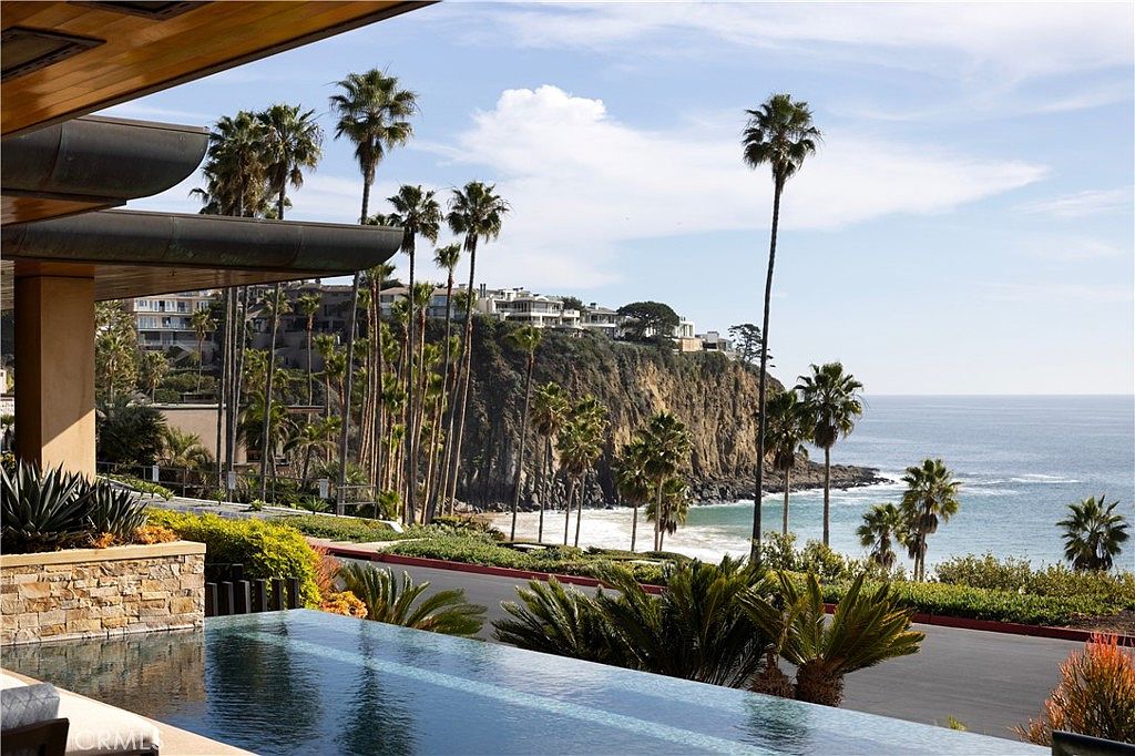 This image showcases an infinity pool overlooking the ocean, framed by palm trees and lush landscaping. The pool's edge blends seamlessly with the horizon, creating a sense of luxury and tranquility. A multi-story building sits atop a cliff in the background, adding to the scenic view.