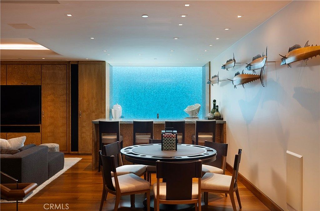 This interior shot showcases a dining room with a unique design. A round poker table with chairs sits in the center, complemented by a bar area with seating against a textured blue wall. The room features hardwood floors, wooden cabinetry, and decorative fish art on the wall, creating a sophisticated and inviting atmosphere.