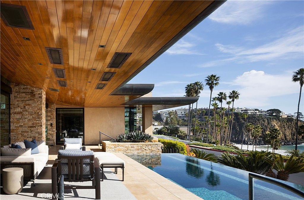 This image showcases a luxurious outdoor patio/deck area with an infinity pool overlooking a stunning coastal view. The space is furnished with modern outdoor seating, complemented by a sleek wooden ceiling and stone accents. The overall impression is one of high-end relaxation and sophisticated outdoor living.