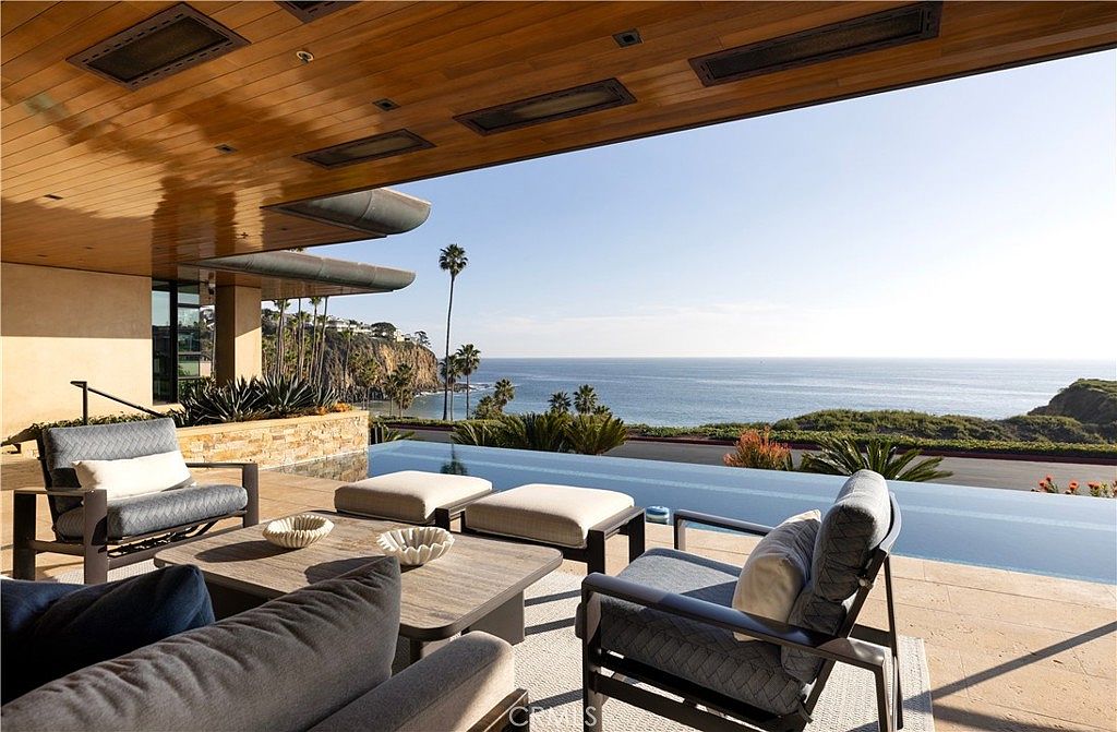 This image showcases a luxurious patio or deck area with comfortable outdoor seating, including chairs, ottomans, and a coffee table. The space overlooks a stunning ocean view, enhanced by an infinity pool that seamlessly blends with the horizon. The wooden ceiling adds a touch of warmth and sophistication to the outdoor living space.