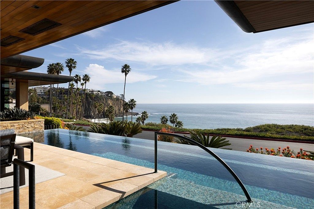This image showcases a luxurious infinity pool overlooking the ocean. The pool area features stone tiling and a sleek, modern design with palm trees and coastal vegetation in the background. The view extends to the ocean and sky, creating a serene and upscale ambiance.